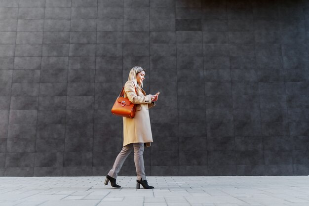 Zijaanzicht van aantrekkelijke Kaukasische blonde vrouw in laag die door grijze muur buiten overgaat en slimme telefoon met behulp van.