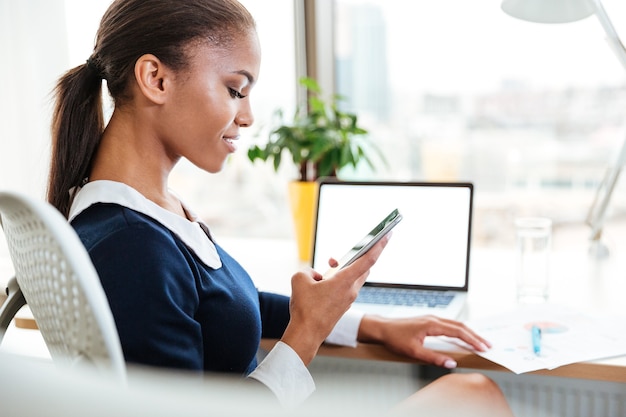 Zijaanzicht van Afrikaanse zakenvrouw in jurk die bij de tafel bij het raam op kantoor zit en naar de telefoon kijkt