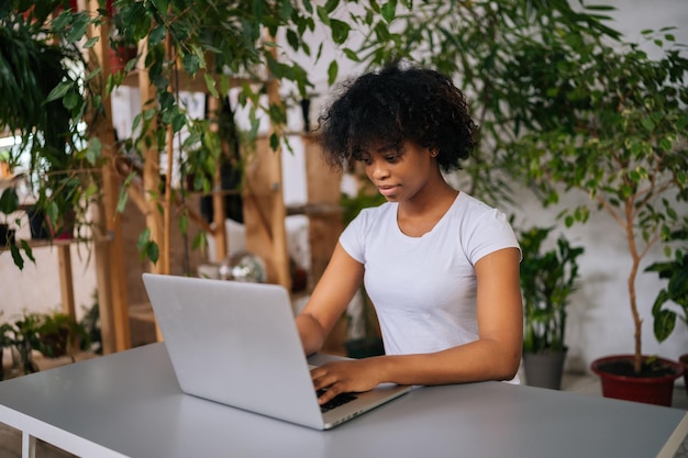 Zijaanzicht van Afro-Amerikaanse zakenvrouw die op afstand werkt aan laptop zittend aan tafel in groene kantoorruimte aan huis met modern biofilie-ontwerp