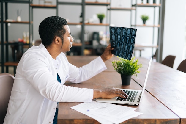 Zijaanzicht van Afro-Amerikaanse zwarte mannelijke arts in witte jas die de geschiedenis van de patiënt analyseert met behulp van MRI-hersenkopscan die op laptop werkt Concept van geneeskunde en gezondheidszorg