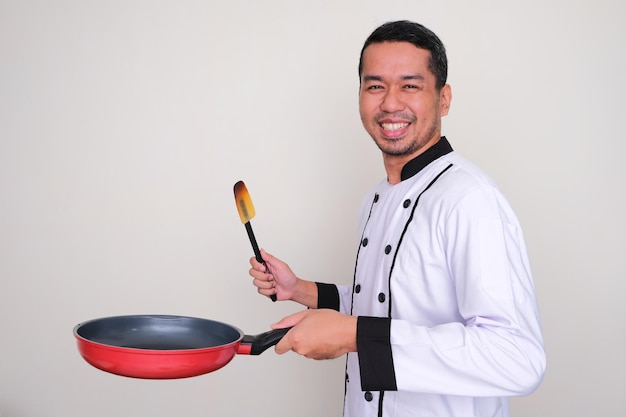 Zijaanzicht van Aziatische chef-kok die blij lacht terwijl hij koekenpan en spatel vasthoudt