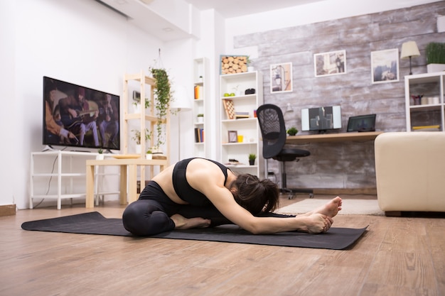 Zijaanzicht van blanke vrouw zittend op de knie naar voren buigen yoga oefening in woonkamer.