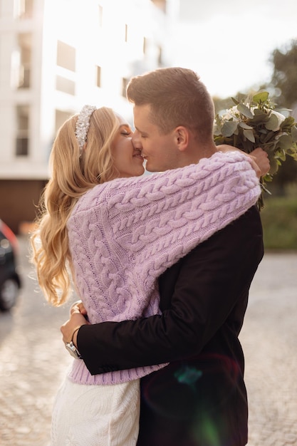 Zijaanzicht van blonde bruid met krullend haar en witte bandeau op hoofd met witte jurk en lila jas met boeket en bruidegom in zwarte jas knuffelen en zoenen buitenshuis op warme zonnige dag