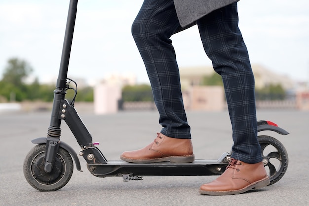 Zijaanzicht van de benen van een zakenman in bruine leren laarzen en een geruite broek die op een elektrische scooter in een stedelijke omgeving op de weg staat