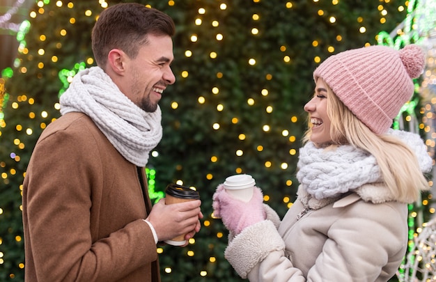 Zijaanzicht van de jonge man en vrouw met afhaalmaaltijden kopjes warme dranken glimlachend en kijken elkaar, terwijl staande in de buurt van de kerstboom op eerste kerstdag