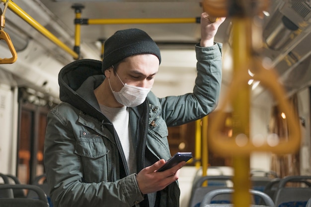 Foto zijaanzicht van de mens met medisch masker dat zijn telefoon op de bus bekijkt