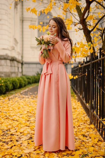 Zijaanzicht van de volledige lengte van een mooie sensuele jonge brunette bruid in roze trouwjurk en permanent in herfst park in de buurt van vintage kathedraal en bruiloft boeket, verticale foto te houden