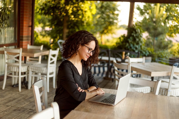 Zijaanzicht van de vrolijke jonge vrouw die aan tafel zit te typen op de laptop op het prachtige terras