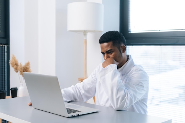 Zijaanzicht van doordachte jonge Afro-Amerikaanse mannelijke beoefenaar arts in wit uniform kijkend naar laptopscherm zittend aan bureau in kliniek kantoorruimte