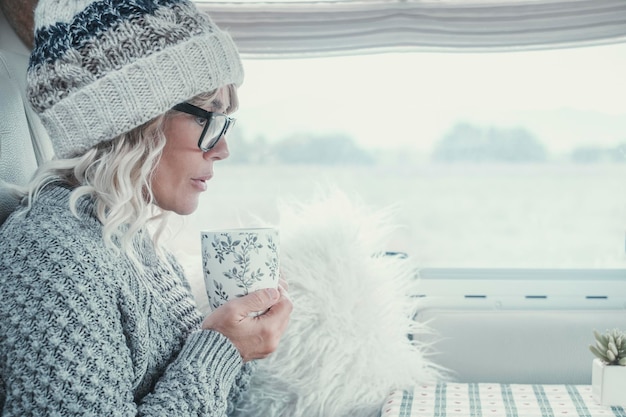 Zijaanzicht van een aantrekkelijke dame met wit haar en kleding die tijdens het winterseizoen drinkt uit een kopje in haar camper helder beeld van een serene vrouw portret van vrouwelijke mensen in de vrije tijd