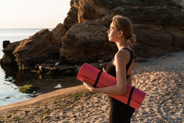 Zijaanzicht van een aantrekkelijke jonge fitnessvrouw met yogamat op het strand, klaar om te trainen