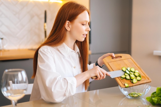 Zijaanzicht van een aantrekkelijke jonge roodharige vrouw die gesneden komkommers in een glazen kom gooit die aan tafel zit