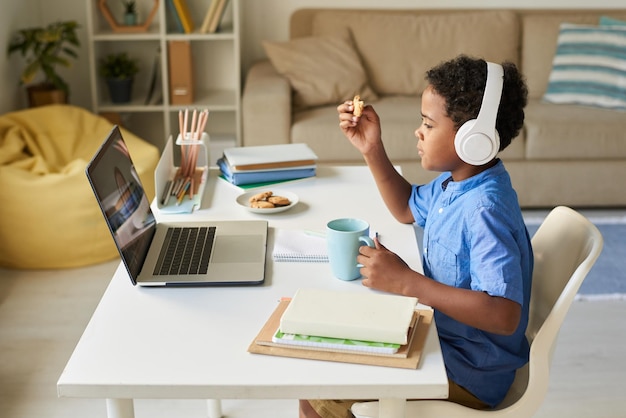 Foto zijaanzicht van een afro-amerikaanse jongen in een draadloze koptelefoon die thuis aan een bureau zit en koekjes eet terwijl