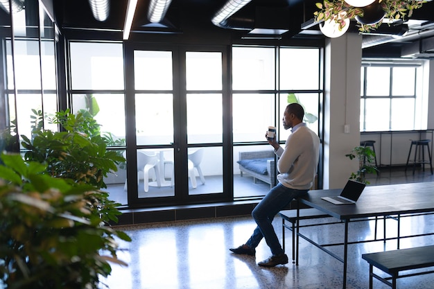 Zijaanzicht van een Afro-Amerikaanse zakenman, werkend in het moderne kantoor, zittend op een bureau en koffie drinkend. Sociale afstand nemen en zelfisolatie tijdens quarantaine-lockdown