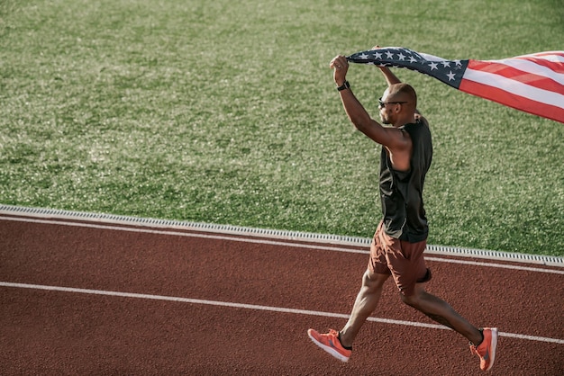 Zijaanzicht van een atleet die op atletiekbaan rent en de Amerikaanse vlag boven het hoofd houdt