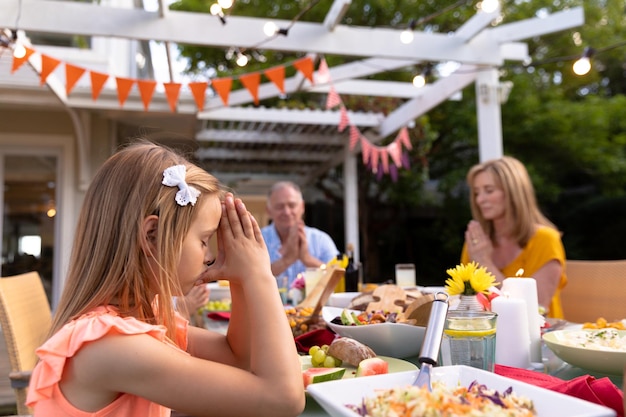 Foto zijaanzicht van een blank meisje dat buiten aan een eettafel zit voor een familiemaaltijd, met gesloten ogen en handen in gebed, genade zeggend voor het eten, haar grootvader en grootmoeder achtergrond