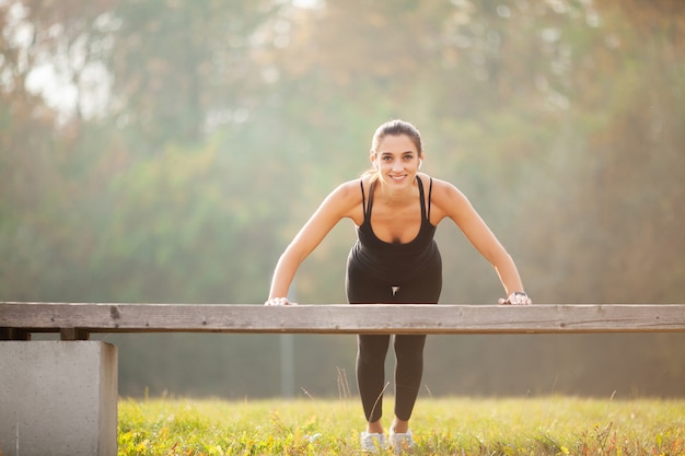 Zijaanzicht van een gemotiveerde Aziatische, portret van mooie sportieve vrouw 20s in sportkleding doet push-ups en luistert naar muziek met bluetooth-oordopjes tijdens training in groen park