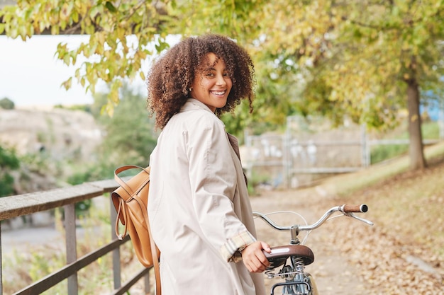 Zijaanzicht van een jonge Afro-Amerikaanse vrouw met krullend haar in een stijlvolle jas die met de fiets staat en naar de camera kijkt in het park