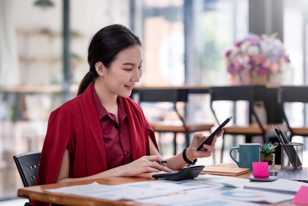 Zijaanzicht van een jonge aziatische zakenvrouw die een smartphone vasthoudt met behulp van een rekenmachine om werkgrafieken te berekenen die op tafel zijn geplaatst.