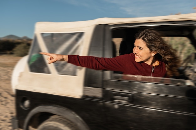 Foto zijaanzicht van een jonge blanke gelukkige vrouw die uit het autoraam kijkt en iets wijst. off-road voertuig in actie.