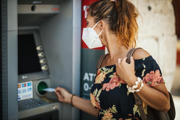 Foto zijaanzicht van een jonge vrouw die een n95 beschermend masker draagt terwijl ze geld aanneemt van een geldautomaat tijdens de covid-19-pandemie.