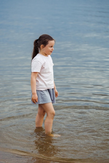 Zijaanzicht van een klein lachend prachtig meisje met lang donker haar met een witte blauwe T-shirtrok die in het water staat