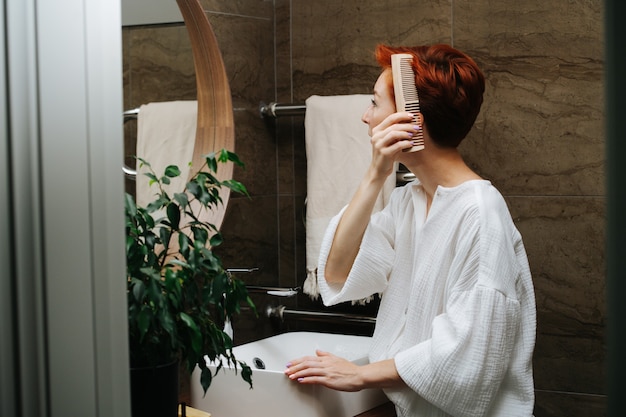 Zijaanzicht van een kortharige volwassen vrouw die haar haar kamt met een houten kam. ze staat voor de spiegel in een badkamer.