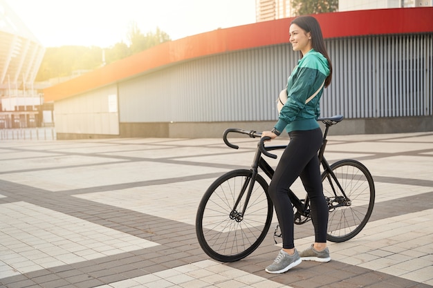 Zijaanzicht van een mooie jonge dame die in de buurt van de fiets staat terwijl ze wegkijkt in de stad. Sport en lifestyle concept
