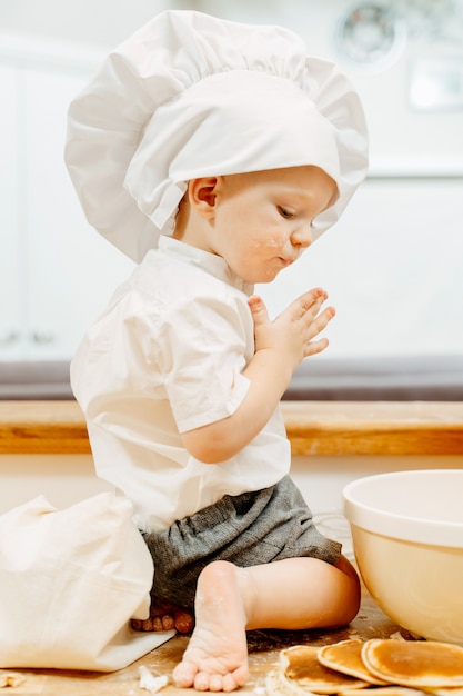 Zijaanzicht van een ondeugende kleine jongen in wit kokkostuum zittend op de keukentafel op blote voeten