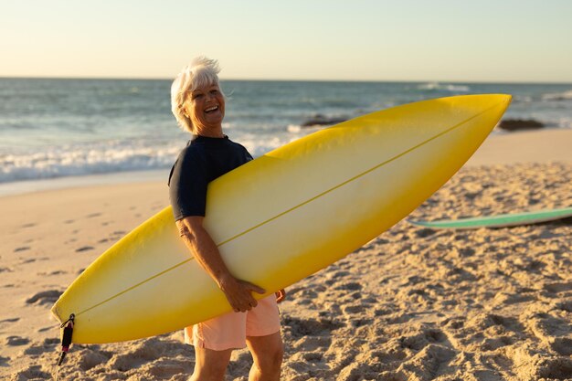 Zijaanzicht van een oudere blanke vrouw op het strand bij zonsondergang, staande op het zand met een surfplank onder haar arm, kijkend naar de camera en glimlachend, met de zee op de achtergrond