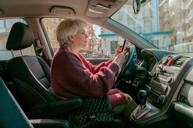 Zijaanzicht van een oudere vrouw die achter het stuur van het motorvoertuig zit en naar haar smartphone staart