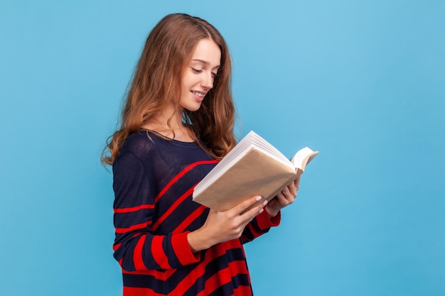 Zijaanzicht van een positieve vrouw die een gestreepte, casual stijltrui draagt, geniet van lezen, in beslag genomen door een spannend plot, het schema in de organisator controleren. Indoor studio-opname geïsoleerd op blauwe achtergrond