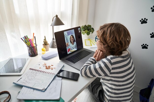 Zijaanzicht van een preteenjongen met krullend haar die aan tafel zit en videochat met een vriend via netbook terwijl hij huiswerk maakt in de kamer