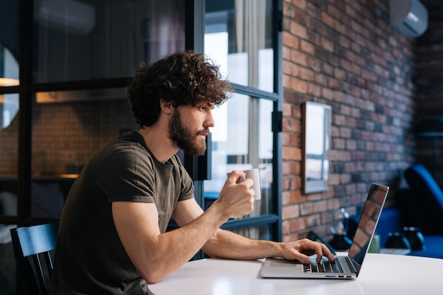 Zijaanzicht van een serieuze jonge zakenman die een kopje in de hand houdt met koffie in de ochtend aan tafel met een laptopcomputer