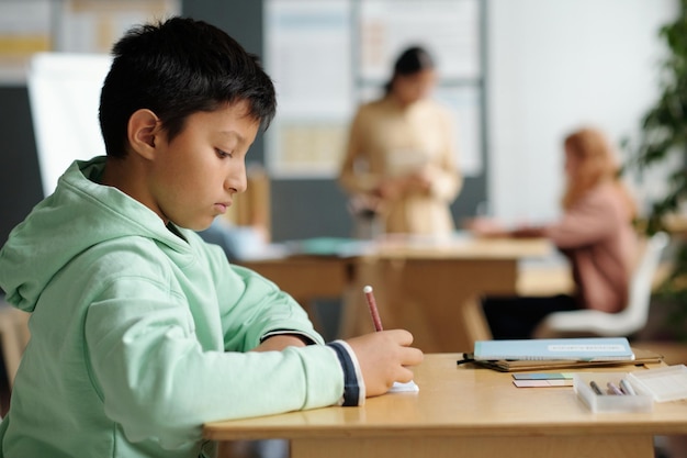 Zijaanzicht van een serieuze multi-etnische schooljongen in vrijetijdskleding die aantekeningen maakt