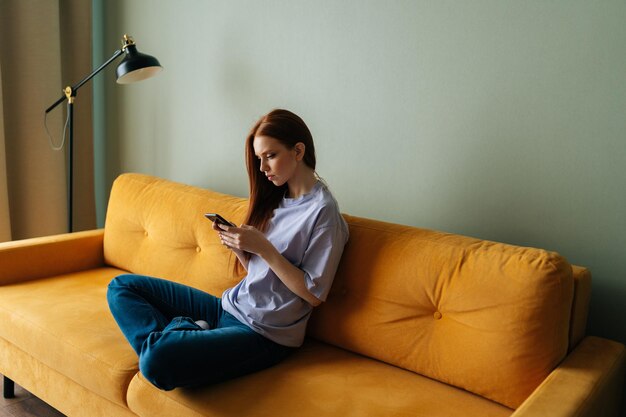 Zijaanzicht van een serieuze roodharige jonge vrouw die naar het smartphonescherm kijkt terwijl ze op een gele bank zit en online chat met behulp van een virtuele app op de mobiele telefoon, winkelen op internet thuis alleen rustend