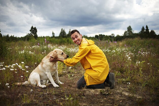 Zijaanzicht van een tevreden jonge man in vrijetijdskleding die lacht terwijl hij op één knie staat voor de labrador retriever in het veld