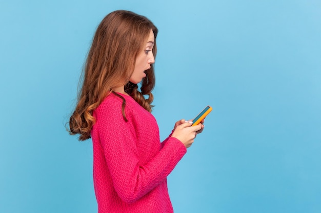 Zijaanzicht van een verbaasde vrouw die een roze trui draagt, geschokt kijkt naar de mobiele telefoon, het laatste nieuws leest op sociale netwerken, applicatie. Indoor studio opname geïsoleerd op blauwe achtergrond.