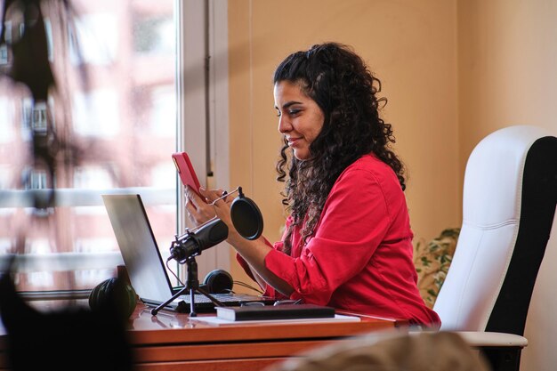 Zijaanzicht van een vrouw die naar het scherm van de mobiele telefoon kijkt terwijl ze thuis werkt met laptop en microfoon