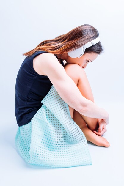 Zijaanzicht van een vrouw die zomerkleren draagt en naar muziek luistert die depressief op de grond zit met een koptelefoon op een witte achtergrond.