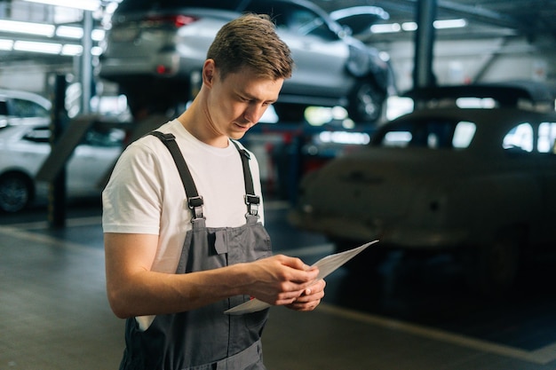 Zijaanzicht van gefocuste knappe jonge monteur man met een uniform lezend klembord in de garage van de autoreparatiewerkplaats met voertuigachtergrond die naar de camera kijkt