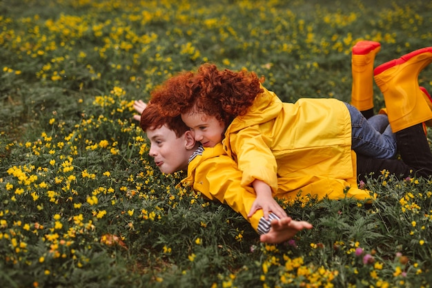 Zijaanzicht van gemberbroer en zus in regenjassen die met uitgestrekte armen op bloeiend gras liggen en doen alsof ze vliegen tijdens het spelen in de natuur op weekenddag