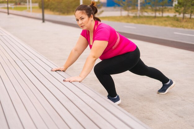 Zijaanzicht van gemotiveerde dikke jonge vrouw die traint en push-ups doet met behulp van een straatbank in het stadspark op de zomerochtend die naar de camera kijkt