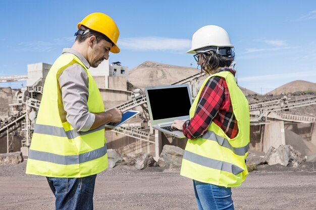 Zijaanzicht van gerichte mannelijke ingenieur met behulp van tablet en vrouwelijke collega typen op laptop terwijl ze samen tegen industriële faciliteit staan