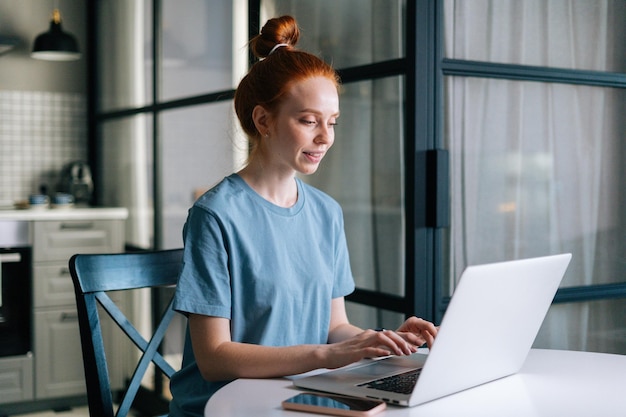 Zijaanzicht van glimlachende roodharige jonge vrouw die aan het typen is op een laptopcomputer die aan tafel zit