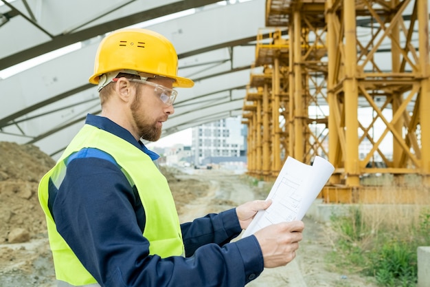 Zijaanzicht van hedendaagse ingenieur die blauwdruk vasthoudt met schets van gebouw