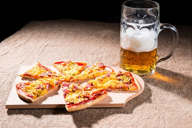 Zijaanzicht van het profiel van mok bier met schuimige kop naast plakjes pizza gerangschikt op houten snijplank op jute gedekt tafeloppervlak