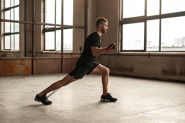 Zijaanzicht van het volledige lichaam van een vastberaden bebaarde man in sportkleding die lunges doet met weerstandstouw tijdens functionele training in de sportschool