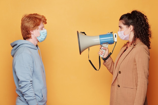 Zijaanzicht van jonge brunette vrouw in beschermend masker speaknig in megafoon terwijl staande voor man in blauwe hoodie