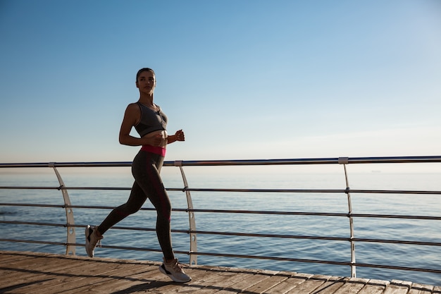 Zijaanzicht van jonge fitness vrouw loopt langs pier.